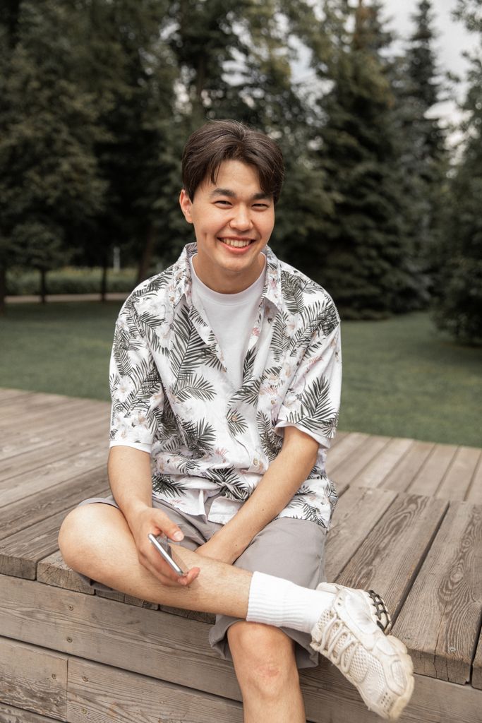 High angle of positive young ethnic man in trendy outfit smiling and looking at camera while relaxing on wooden bench in green park